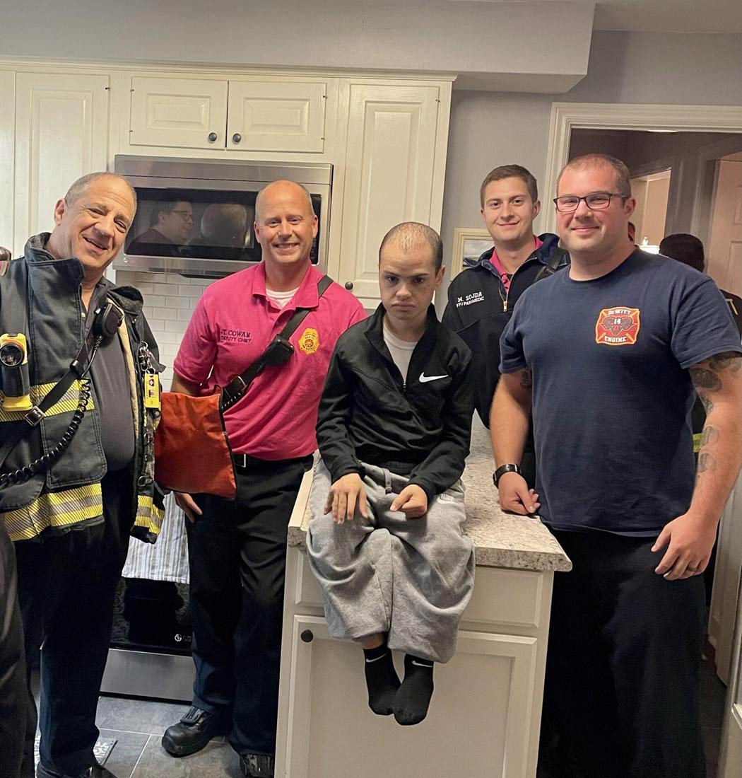 A young man sits on a kitchen counter surrounded by the smiling firefighters.
